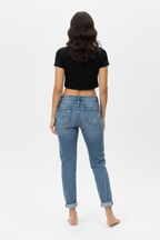 Back full-body shot of a female model wearing paint-splatter boyfriend jeans and a black short-sleeved crop top, barefoot, standing against a white background.