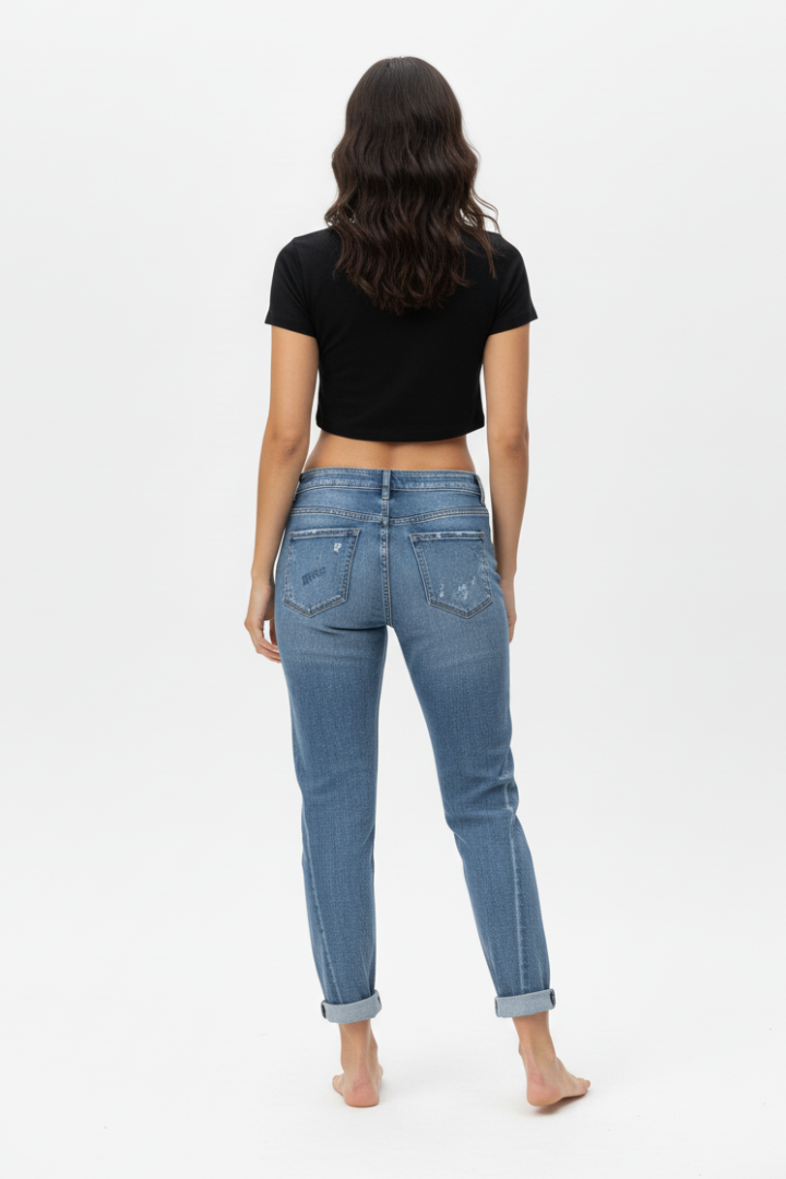 Back full-body shot of a female model wearing paint-splatter boyfriend jeans and a black short-sleeved crop top, barefoot, standing against a white background.