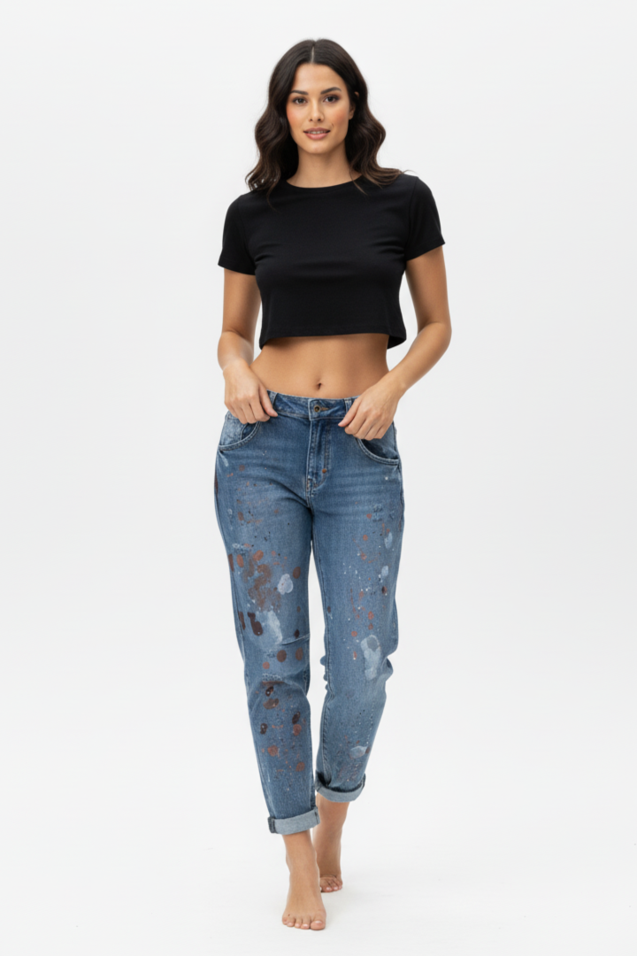 Full-body shot of a female model wearing paint-splatter boyfriend jeans and a black short-sleeved crop top, barefoot, standing against a white background.
