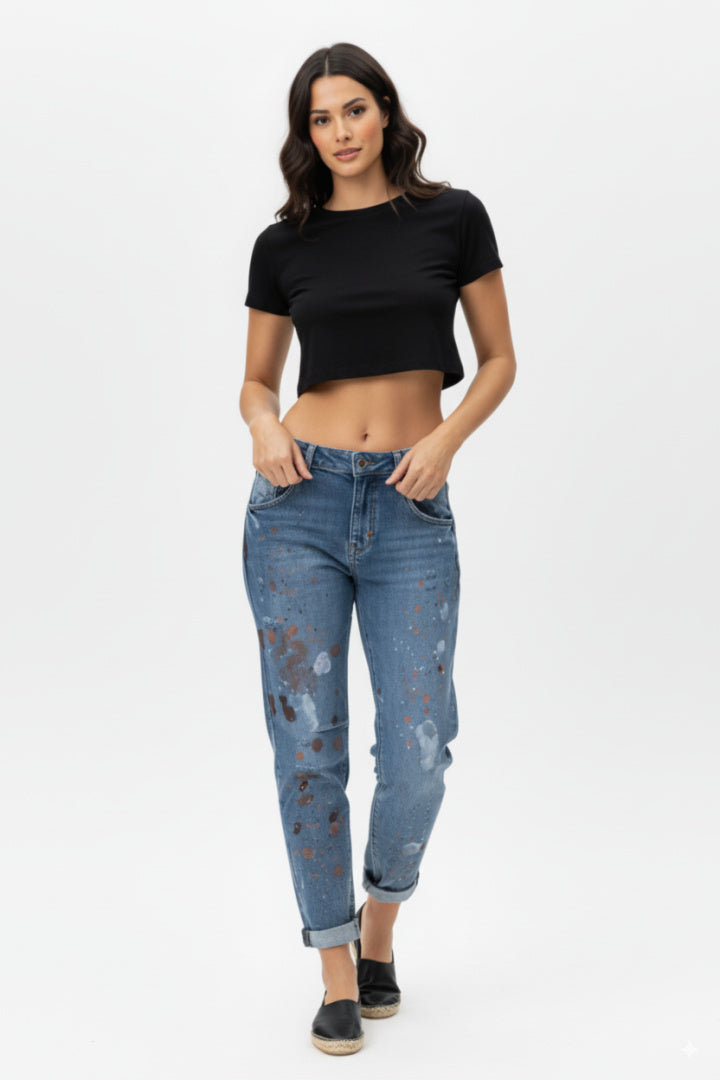 Side full-body shot of a female model wearing paint-splatter boyfriend jeans and a black short-sleeved crop top, barefoot, standing against a white background.