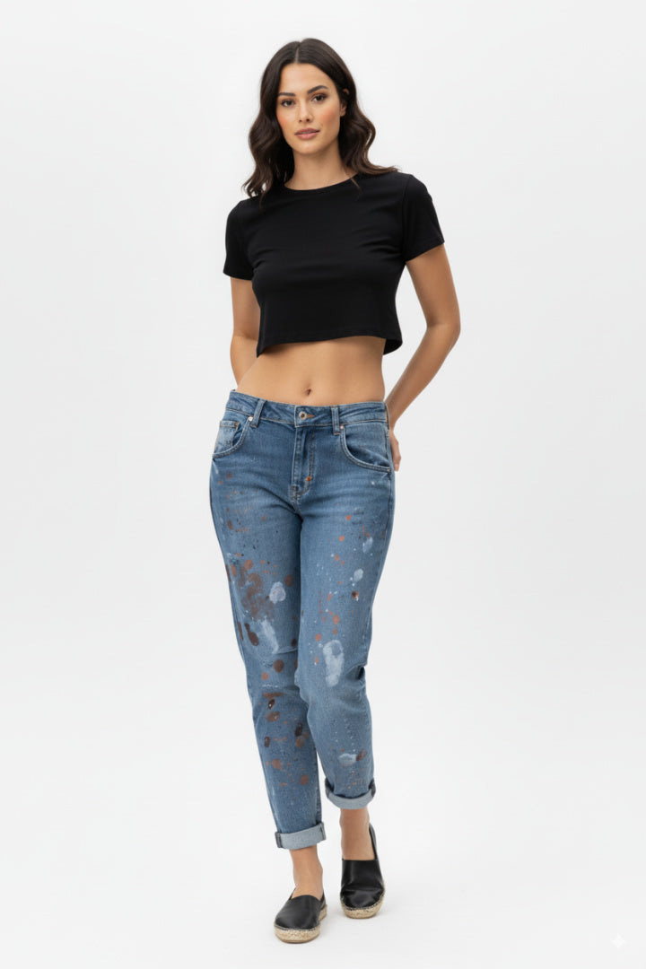 Side hand full-body shot of a female model wearing paint-splatter boyfriend jeans and a black short-sleeved crop top, barefoot, standing against a white background.