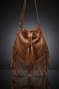 A close-up, studio shot of a rustic brown genuine leather hobo bag. The bag has a full fringe trim, a single shoulder strap, and a braided basketweave pattern on the front. Two decorative tassels hang from the drawstring closure. The bag's reflection is visible on a dark, glossy surface.