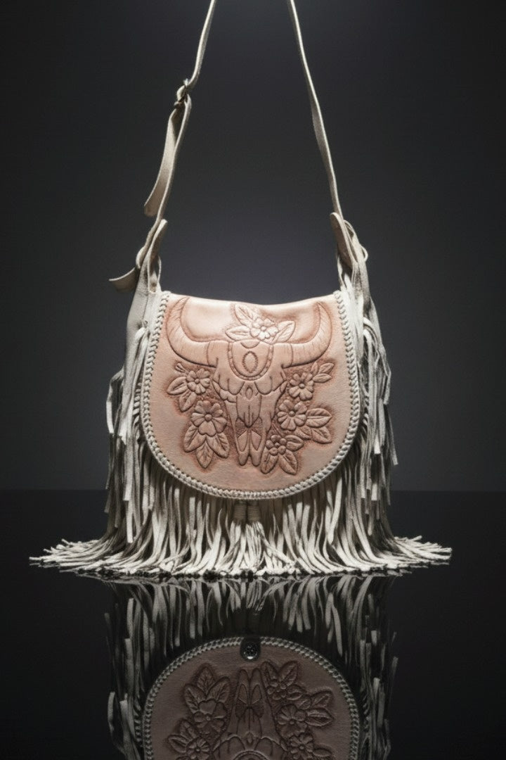 Stylish bohemian handbag in beige leather with long fringe detailing and a light brown flap featuring an embossed bull’s head and floral design, displayed against a dark reflective background.