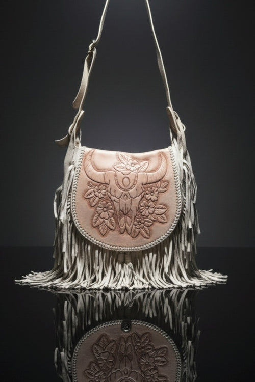 Stylish bohemian handbag in beige leather with long fringe detailing and a light brown flap featuring an embossed bull’s head and floral design, displayed against a dark reflective background.
