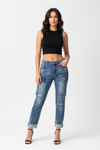 Full-body shot of a female model wearing distressed patchwork paint-splatter boyfriend jeans with a black ribbed sleeveless crop top and black high-heeled sandals, standing against a white background.