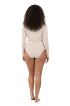 A full-length, studio shot of a smiling woman with curly hair wearing a simple, skin-tone or beige long-sleeve bodysuit with a mock neck.  Back