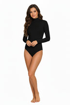 A full-length, studio shot of a smiling woman with wave hair wearing a simple, black long-sleeve bodysuit with a mock neck. 