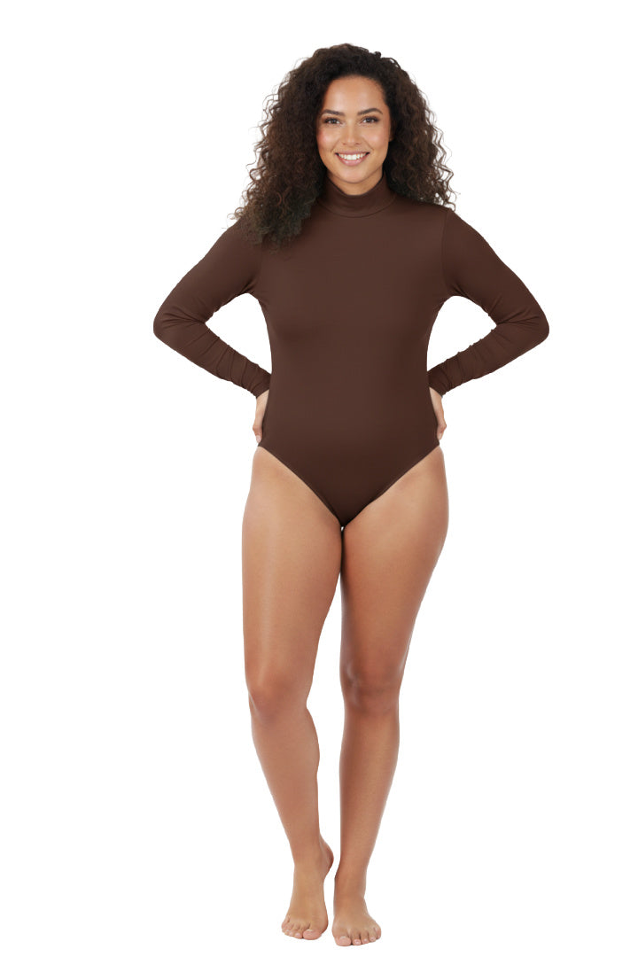 A full-length, studio shot of a smiling woman with curly hair wearing a simple, brown long-sleeve bodysuit with a mock neck. She is standing with her hands on her hips against a white background.