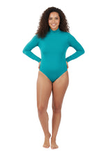 A full-length, studio shot of a smiling woman with curly hair wearing a simple, turquoise long-sleeve bodysuit with a mock neck. She is standing with her hands on her hips against a white background.