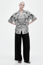 A full-length studio shot of a model wearing the "Shiva Lover" short-sleeve, open-collar shirt with a complex black and white art pattern. The shirt is paired with wide-leg black trousers.