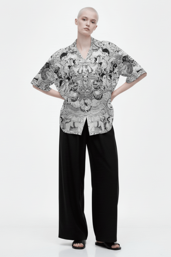 A full-length studio shot of a model wearing the "Shiva Lover" short-sleeve, open-collar shirt with a complex black and white art pattern. The shirt is paired with wide-leg black trousers.