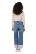 A model wearing a pair of light blue, high-rise, wide-leg jeans with a contrasting wide cuff at the ankle. The jeans have a classic five-pocket design and are paired with a beige top and white shoes. The model is standing in a studio setting. Back