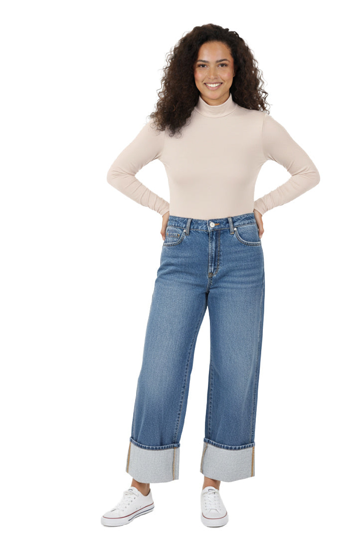 A model wearing a pair of light blue, high-rise, wide-leg jeans with a contrasting wide cuff at the ankle. The jeans have a classic five-pocket design and are paired with a beige top and white shoes. The model is standing in a studio setting. Front